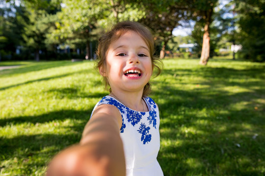 Beautiful kid taking selfie in green summer park, laughing, having fun. Warm sunny weather, healthy child, happiness, freedom, emotions. Cute girl reaching hand to camera. 