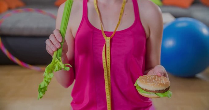 A girl can't decide whether she shall choose to a stalk of celery or a hamburger. She taps a burger with the celery.
