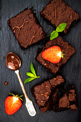 Homemade dark chocolate brownies decorated with strawberries and mint leaves over black slate background, top view