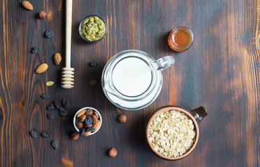 Obraz na płótnie Canvas Ingredients for healthy breakfast - bowls with oatmeal, almond and hazel nuts, raisins and pumpkin seed, honey jar, honey spoon, milk jugful on rustic wooden background, top view