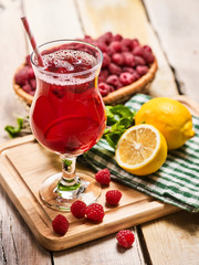 On wooden boards is ice cold cocktail glass with red raspberry lemon cocktail . Alcohol country cocktail beverage number hundred twenty eight with raspberry clay bowl. Cocktail on checkered napkin.
