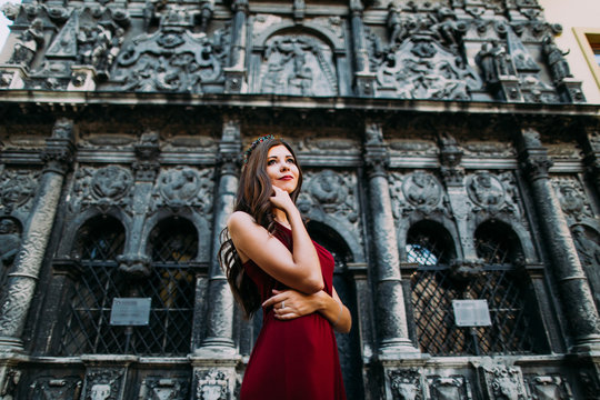 Cheerful young brunette in a red dress posing with old Lviv cathedral on background