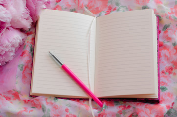 Open notebook and pen on the background of pink floral fabric next to flowers