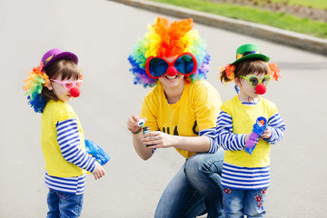 Young woman and two charming daughters in clown costume blowing bubbles outdoors at summer day