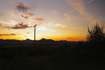 Fototapeta premium romantic sunset in the region Ceske stredohori in czech landscape with fields, pylons and mountains Milesovka and Lovos
