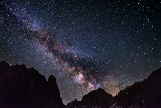 The colorful glowing core of the Milky Way and the starry sky captured at high altitude in summertime on the Alps. Scenic snowcapped mountain silhouette.