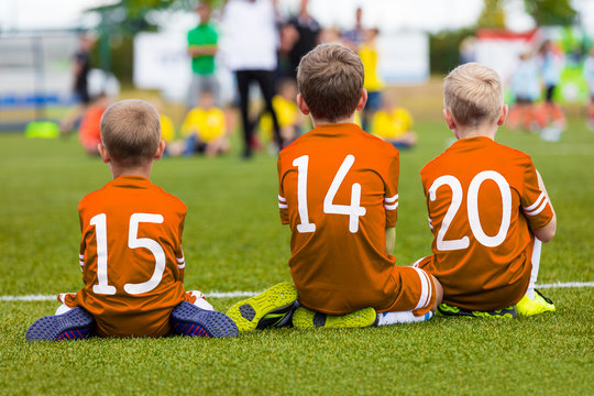 Kids of young soccer football team. Boys in orange shirts as reserve players sitting on football pitch and watching soccer match.