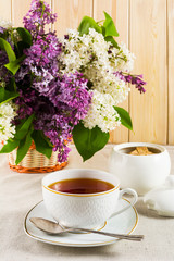 Cup of tea and branch of lilac flower in wicker basket on linen