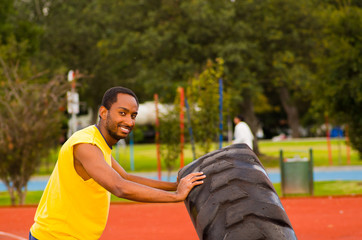 Man wearing yellow shirt and blue shorts lifting large tractor tire during strength excercise, outdoors training facility with orange athletic surface, green trees background