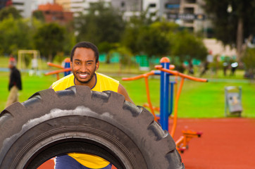 Obraz na płótnie Canvas Man wearing yellow shirt and blue shorts lifting large tractor tire during strength excercise, outdoors training facility with orange athletic surface, green trees background