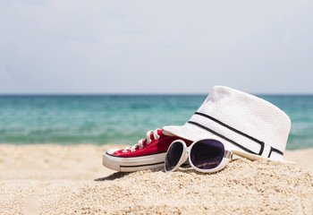 Close up of white hat, sunglasses and red sneakers on the beautiful sand beach as summertime concept. Beach accessories concept