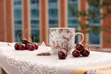 Чай в большой кружке с рисунком веток дерева вишни. Рядом лежит чайный пакетик и ягоды. Все предметы расположены на фоне окон здания на балконе.