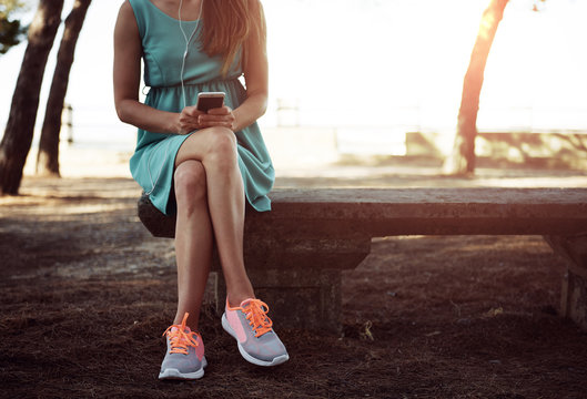 Hipster girl in green dress and sneakers holding smart phone and listening music in earphones in the park. Concept of modern technology and lifestyle. 
