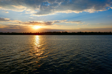 Orange setting sun over water surface with ripples and tree tops on far coast