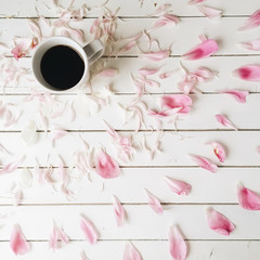 Cup of black coffee and pink peony petals on white wooden background. Overhead view