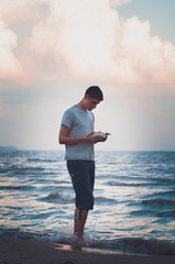 teenage boy reading a book while standing in the waters edge at a beach