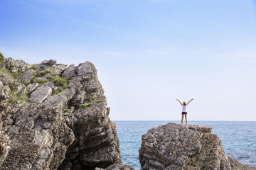 Happy woman with hands up standing on cliff over sea and islands at sunset.
