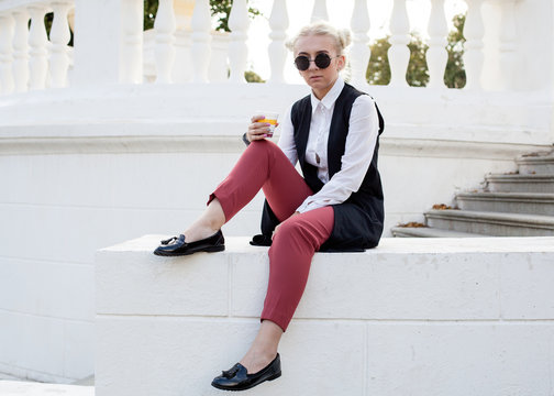 Trendy young woman in sunglasses sitting on the parapet with a cup of coffee