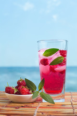 Raspberry lemonade with mint and ice on wooden table with sea on background. Healthy fresh summer drink with berries.