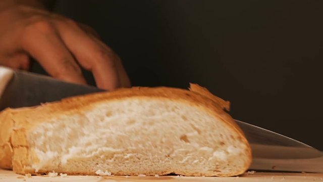Baker is cutting slice of his freshly baked bread with kitchen knife on a wooden surface
