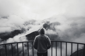 View from behind of a man standing at view point looking to beautiful landscape with foggy mountains in the distance. Black and white photo. - 115754768