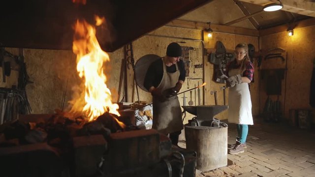 Forge master and journeyman woman unfetters metal rod.