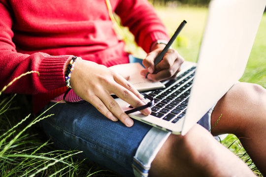 Midsection of university student writing in book while using laptop at park