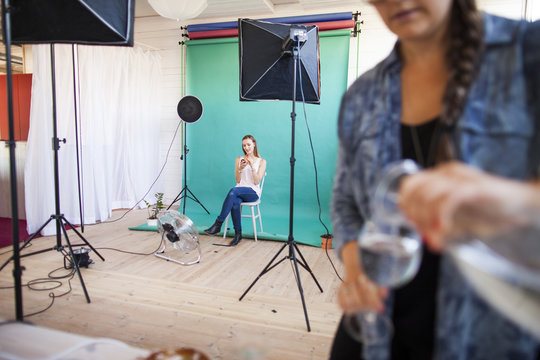 Photo assistant pouring water in glass while fashion model getting dressed in background at studio