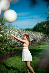 A charming girl holding white balloons looking sadly at them, ready to let them go. Blossoming garden and spring sky on the background