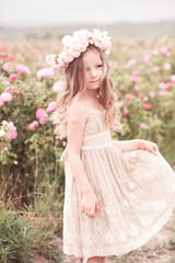 Smiling kid girl 4-5 year old posing in rose garden outdoors. Wearing stylish dress and wreath with flowers. Summer portrait