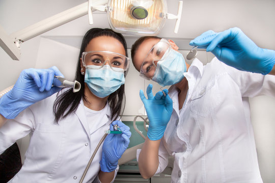 Female doctor dentist and her assistant in masks and white uniforms holding in their hands dental mirror, probe and other equipment, looking at camera and smiling