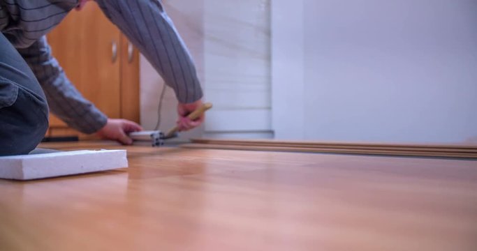 A man is using a hammer to check if all sides are even. He and his friend are laying wood panels to build a new floor. Close-up shot.
