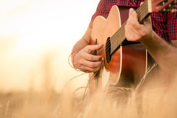 Close up of male hands playing acoustic guitar on the wheat field at the sunset. Retro, music,...