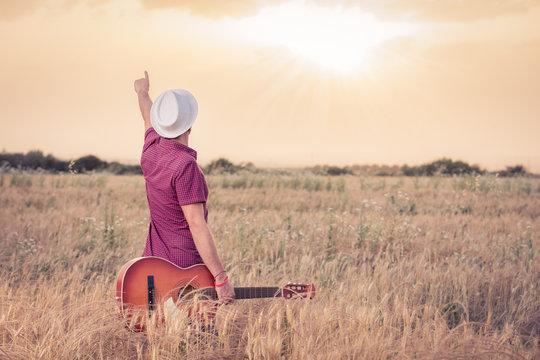 Young retro styled man holding acoustic guitar in wheat field and looking at sun to find inspiration for the next song. Music, art and lifestyle concepts.  