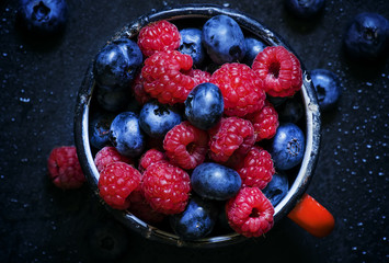 Raspberries and blueberries in old enamel mugs, dark food backgr