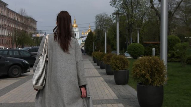 Back view of a stylish female tourist in gray coat and with striped bag admires of a beautiful architectural building during walking in foreign city