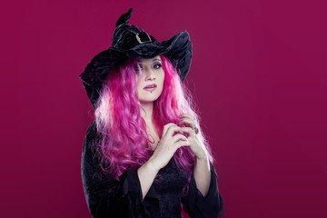 Attractive woman in witches hat and costume with red hair performs magic on a pink background. Halloween, horror theme.