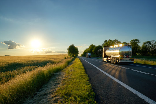 Trucks with chrome tank driving on asphalt road along the corn field at sunset. The landscape and the road are mirrored on a silver tanker.