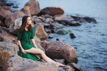 Cute girl in green dress sitting barefoot on a large rock on the