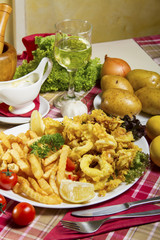 Serving of breaded and deep-fried calamari and fries on a white plate served on a table with fresh vegetables,  dressing sauce, lemon and a glass of white wine.
