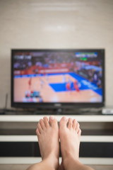 man laying down on a sofa at home watching basket match on tv