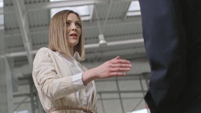 Low angle view of beautiful young woman standing, talking with male colleague and giving him handshake in business center