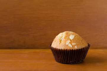 A piece of delicious banana cup cake topped with sliced almond in dark brown paper cup over orange brown teakwood table with wooden background. Fresh baked cup cake with space, selective focus