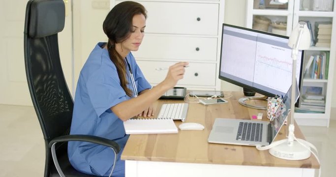 Young female doctor working at her desk in the office writing notes as she reads online information on her patients or current medication