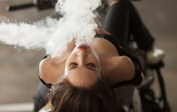 Young sexy woman sitting on motorcycle and smoking ( vaping ) e-cigarette.
