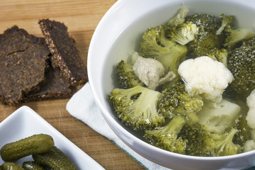 White bowl of soup with chicken and vegetables, with bread and cucumbers on the wooden background