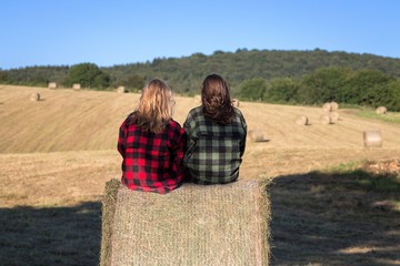 au milieu d'un champ de récolte, deux jeunes filles sont assises de dos sur un rouleau de paille