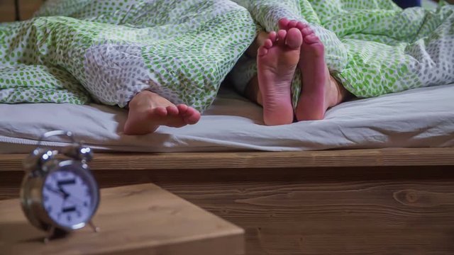Lady's feet are slowly touching her husband's feet in bed. They are lying down. Alarm clock is standing on the bedside table.
