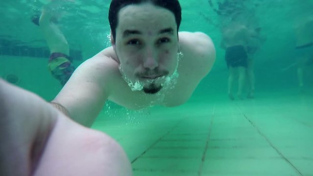 A young man is swimming under water with a camera in his hand. He is having no googles on. Close-up shot.
