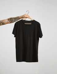 Tattooed biker hand holds hang with blank black t-shirt from premium thin cotton, isolated on white mockup